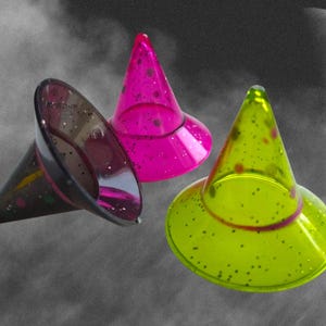 May include: Three colorful witch hats in the image. One is black, one is bright pink, and the other is lime green. Each hat has a conical shape with a wide brim and is decorated with small, dark spots. The background is a cloudy, grayscale scene with a moon.