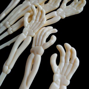 May include: Off-white plastic skeleton hand-shaped serving forks. The forks have a detailed bone structure and are arranged against a black background. The forks are ideal for Halloween or themed parties.