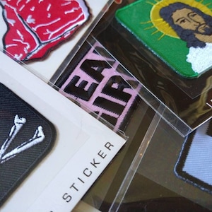 May include: Assortment of iron-on patches featuring various designs, including a steak, a skull and crossbones, a religious figure, and a checkered pattern. The patches are packaged in clear plastic bags with labels that say "Iron-On" and "Sticker".