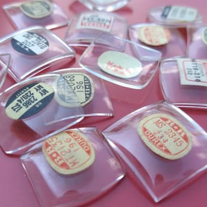 May include: A collection of clear, square-shaped glass objects with rounded edges, each containing a small, aged paper label. The labels feature text and numbers, some with the words "Made U.S.A." and "DUREX". The objects are scattered on a pink surface.