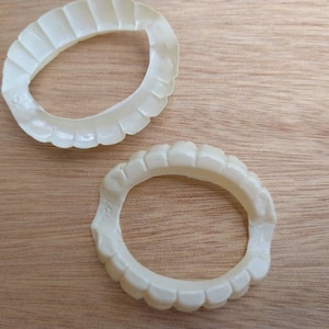 May include: Two off-white, plastic vampire teeth sets, each with a scalloped edge, rest on a light brown wooden surface. The teeth are designed to fit over the natural teeth, creating a spooky effect.