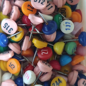 May include: A collection of colorful push pins with various numbers printed on them. The pins have round, plastic heads in shades of pink, yellow, orange, blue, green, and red. The numbers are printed in black ink.