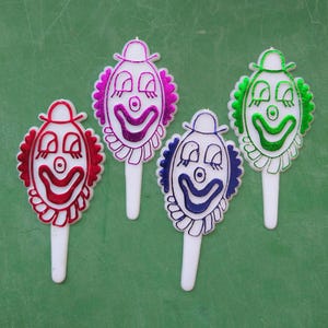 May include: Four colorful clown face cake toppers with white sticks. The clown faces are pink, green, red, and blue with black eyes and a white nose.