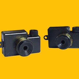 May include: Two black plastic toy cameras with gold accents. The cameras have a small lens and a button on the top.