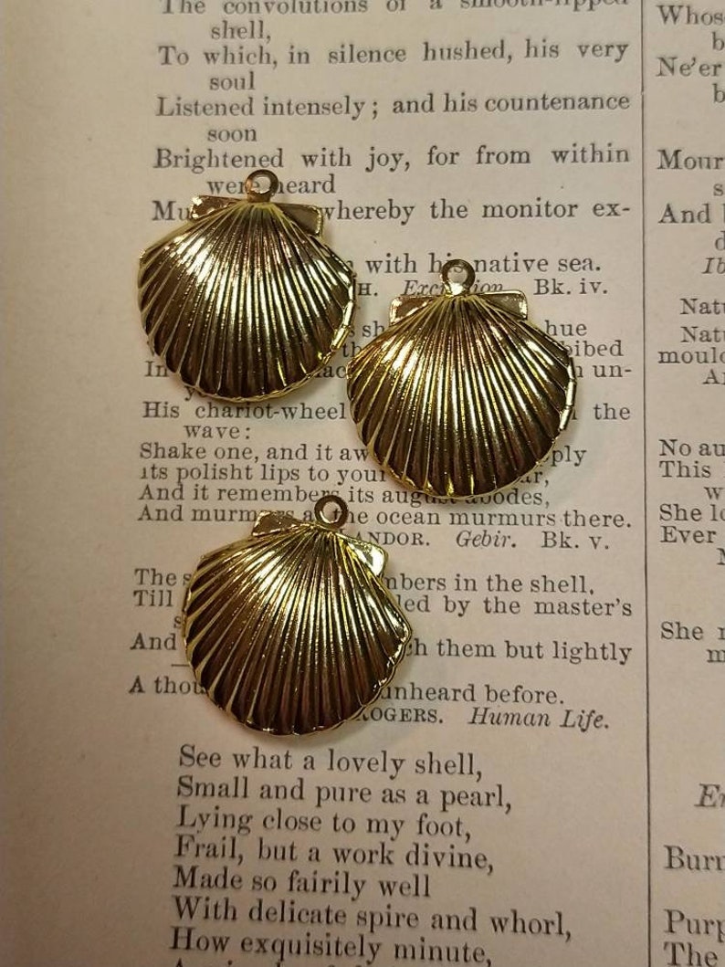 May include: Three gold-colored seashell charms with a textured surface, each with a small loop for attaching to jewelry or other crafts. The charms are displayed on a vintage book page with printed text.