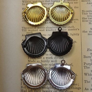 May include: Close-up of four seashell-shaped lockets in gold, black, and silver. Each locket is open, revealing the interior design. The lockets are on a vintage book page with printed text.