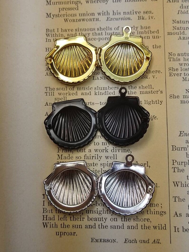 May include: Close-up of four seashell-shaped lockets in gold, black, and silver. Each locket is open, revealing the interior design. The lockets are on a vintage book page with printed text.