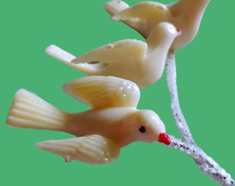 May include: Three cream-colored bird figurines with small red beaks, each perched on a textured, silver-white branch. The birds are depicted in various poses, with one bird shown in flight with wings spread, while the others are resting on the branch.