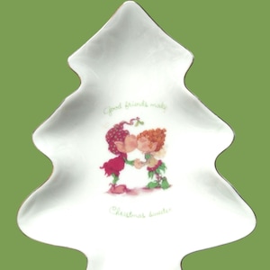 May include: White ceramic Christmas tree-shaped plate with a gold trim. The plate features a cartoon illustration of two figures kissing, with the text "Good friends make Christmas sweeter."