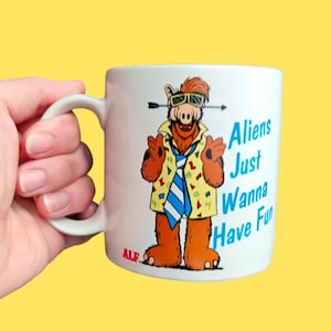 May include: A white ceramic mug featuring a graphic of the character ALF wearing yellow patterned clothing, a blue and white striped tie, and sunglasses. The text "Aliens Just Wanna Have Fun" is printed in blue, with the red "ALF" logo below.