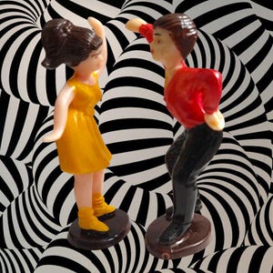 May include: Two small figurines of a dancing couple. The woman wears a yellow dress and boots, while the man is in a red shirt and black pants. They stand on brown circular bases against a black and white striped background.