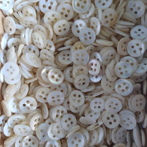May include: A close-up shot of a pile of off-white, pearlescent buttons. Each button has four holes and a slightly raised edge. The buttons are scattered, creating a textured surface. The buttons are likely made of mother-of-pearl.