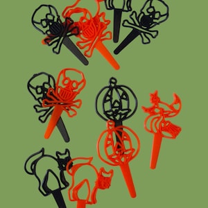May include: A collection of Halloween-themed decorative picks in black and orange. The picks feature designs of skulls with crossbones, black cats, pumpkins, and witches. The picks have a stick for easy insertion into food or decorations.