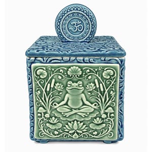 May include: A square ceramic jar with a blue lid featuring a circular Om symbol. The front panel is light green and displays a relief of a frog sitting in a meditative pose, surrounded by detailed botanical illustrations of lotus flowers and reeds.