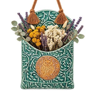 May include: A teal ceramic wall pocket with an embossed vine pattern and a central circular medallion featuring an owl. The pocket is filled with dried lavender, yellow yarrow, white flowers, and eucalyptus, and hangs from two brown rope tassels.