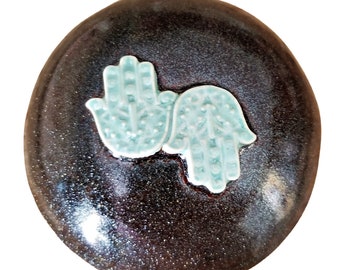 May include: A dark brown ceramic oval with two raised, light blue Hamsa hands. The hands are facing each other and have intricate designs.
