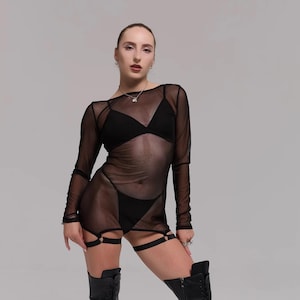 May include: Black lingerie set featuring a sheer long-sleeve dress, a bra, and high-waisted bottoms. The outfit includes garter straps and is paired with knee-high, lace-up boots with high heels. The model is wearing a necklace.