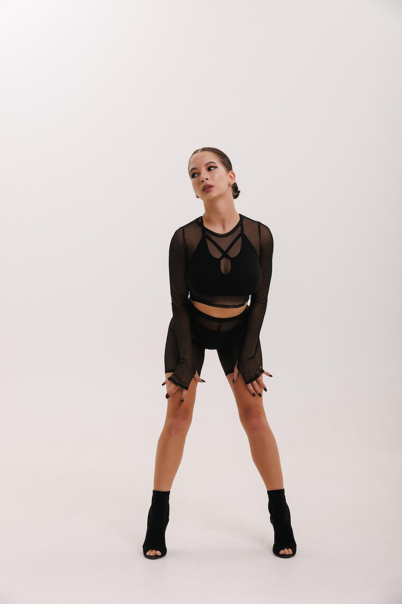 May include: A woman wearing a black mesh long-sleeved top and matching shorts. She is standing with her legs slightly bent and her arms at her sides. She is wearing black heeled boots.