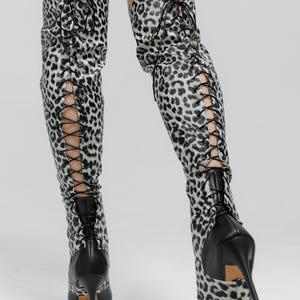 May include: Thigh-high boots with a black and white leopard print pattern. The boots feature a lace-up design on the sides and a pointed toe. The heels are black and have a stiletto design.