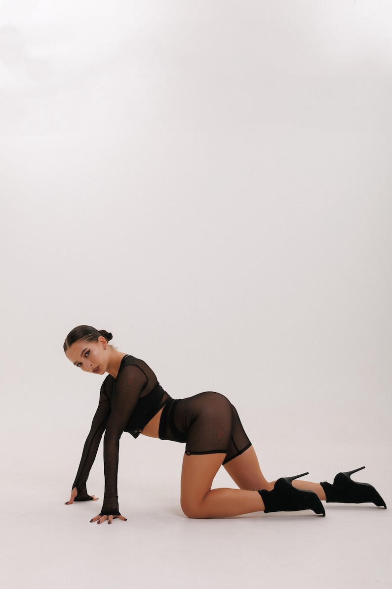 May include: A woman wearing a black mesh crop top and shorts poses on a white background. She is wearing black heeled boots.