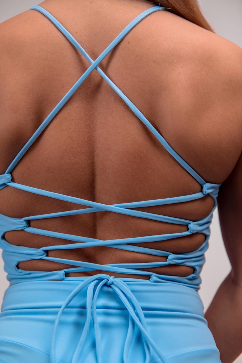 May include: Light blue top with criss-cross back straps. The straps create a lattice design across the back. The top is a solid light blue color and appears to be made of a smooth, possibly stretchy material. The straps tie at the bottom.