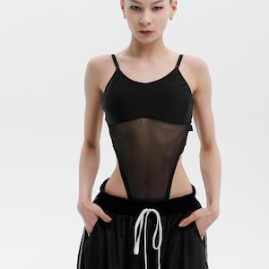 Può includere: Body nero con un pannello in rete trasparente e spalline sottili. Il body è abbinato a pantaloni sportivi neri con coulisse e dettagli bianchi. La modella è in piedi davanti a uno sfondo bianco.