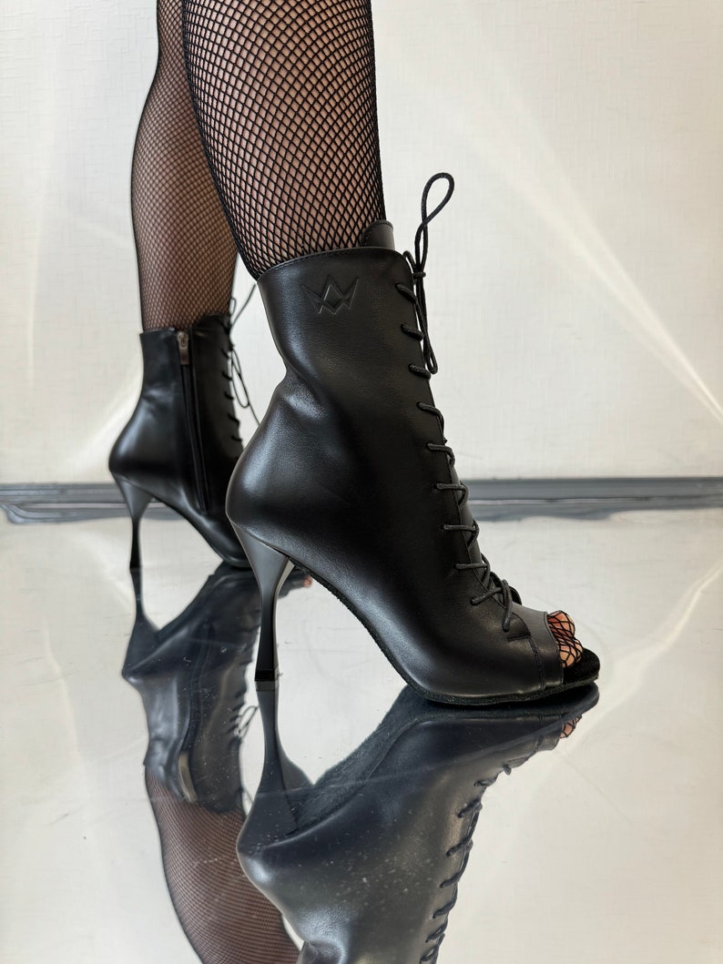 Puede incluir: Botas negras de cuero con punta abierta, detalle de cordones y cremallera lateral. Las botas tienen un tac&oacute;n de aguja y una punta puntiaguda.