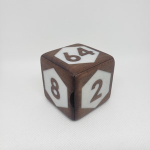 Può includere: Un dado di legno a più facce, con facce intarsiate bianche che mostrano i numeri 2, 4, 8 e 64. Il dado è in legno marrone scuro e poggia su una superficie bianca. Un dado unico per un gioco da tavolo.