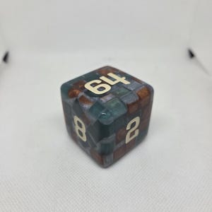 May include: A large, multi-sided die with a mosaic pattern in shades of brown, green, and blue. The numbers 64, 8, and 2 are visible in a cream color. The die is a unique game piece.