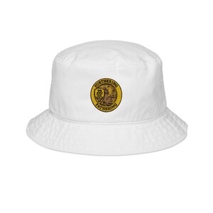 Puede incluir: Sombrero de pescador blanco con un emblema circular en la parte delantera. El emblema presenta las palabras "FURTHERING" y "FATHERING" alrededor de un diseño central. El sombrero está hecho de un material suave y tiene un ala ancha.
