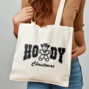 May include: A white canvas tote bag with black text that reads "Howdy Christmas" and a gingerbread man wearing a cowboy hat.
