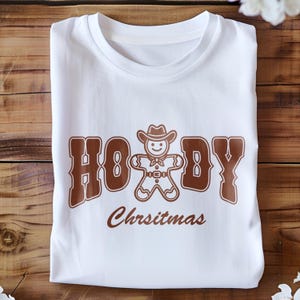 May include: White t-shirt with a brown graphic of a gingerbread man wearing a cowboy hat and the text "Howdy Christmas".