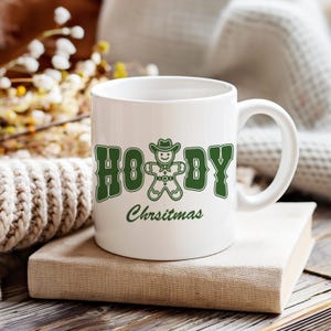 May include: White ceramic mug with a green design that says "Howdy Christmas" and features a gingerbread man wearing a cowboy hat.