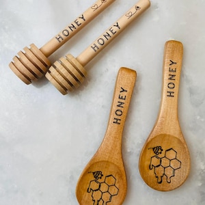May include: Wooden honey dippers and spoons. Two honey dippers with the word "HONEY" and a heart and flower design. Two spoons with the word "HONEY" and a honeycomb design. All items are made of light-colored wood.