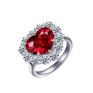 Może przedstawiać: Srebrny pierścionek z czerwonym kamieniem w kształcie serca, otoczonym aureolą mniejszych, przezroczystych kamieni. Pierścionek ma polerowaną srebrną obrączkę i szczegółowe osadzenie.