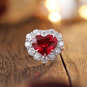 May include: A heart-shaped ruby ring with a halo of white diamonds. The ring features a red gemstone surrounded by smaller, round, clear diamonds. The ring is set on a silver band, with a blurred wooden background.