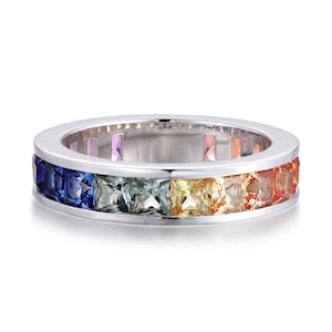 Puede incluir: Un anillo plateado con una fila de piedras preciosas cuadradas en colores del arcoíris. Las piedras están dispuestas en secuencia, desde el azul hasta el rojo. El anillo tiene una superficie pulida y reflectante.