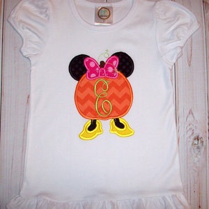 May include: White t-shirt with a pumpkin-shaped Minnie Mouse design. The pumpkin is orange with a chevron pattern and has a green letter "E" in the center. Minnie Mouse has black ears, a pink bow, and yellow shoes.