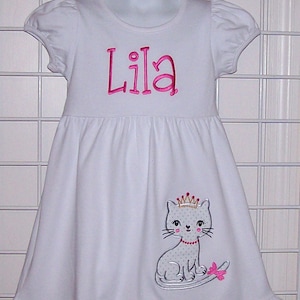 May include: White cotton dress with a pink embroidered name "Lila" and a gray cat with a crown and pink bow.