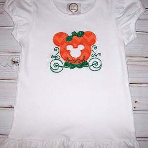 May include: White short-sleeved t-shirt with a pumpkin-shaped Mickey Mouse head design in orange and green. The design features a green bow and a pumpkin carriage.