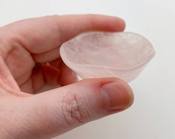 May include: A small, light pink, heart-shaped dish. The dish is made of a translucent material, possibly a crystal or stone. The dish is held in a hand against a white background.