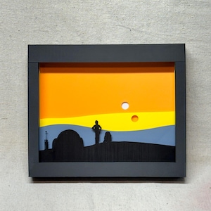 May include: A framed silhouette artwork depicting a desert landscape with a single figure standing in the foreground. The sky is a gradient of orange, yellow, and blue, with two white circles representing the twin suns. The artwork is framed in black.