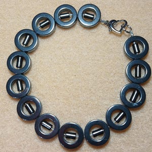 May include: A bracelet composed of dark gray, metallic-looking circular ring beads, each containing a small, cylindrical bead suspended in the center. The piece is secured with a silver-toned metal lobster claw clasp and a circular jump ring.