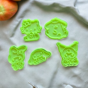 Pode incluir: Cinco cortadores de biscoitos de plástico verde em forma de personagens de desenhos animados. Os cortadores estão sobre um fundo de tecido branco.