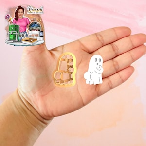 May include: A hand displays two ghost-shaped items. One is a gold-coloured cookie cutter, and the other is a white ghost-shaped object. The cookie cutter has a detailed design, while the other ghost is a simple outline.