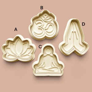 May include: Four beige cookie cutters in yoga and meditation shapes. The shapes include a lotus flower, the Om symbol, a seated figure in meditation, and hands in prayer. The cutters are arranged on a light brown surface.