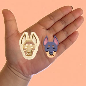 May include: A close-up of a hand holding two dog-shaped cookie cutters. One cutter is beige and the other is purple with a brown nose and ears.
