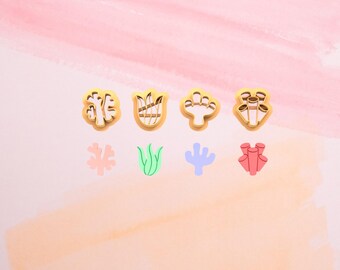 May include: An assortment of gold-colored cookie cutters in various shapes, including coral, a tulip, and a cactus. Below the cutters are colorful, smaller cutouts of the same shapes. The background is a gradient of pink and peach.