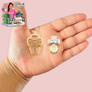 May include: Two cross-shaped cookie cutters. One is a gold-colored outline of a cross with a baby and stars. The other is a cross-shaped pin with a baby, star, and ribbon design. The items are held in a hand.
