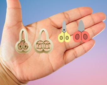 May include: A variety of craft tools are displayed, including two beige plastic cookie cutters shaped like scissors and two pairs of small scissors. One pair is yellow and the other is red, both with silver blades.